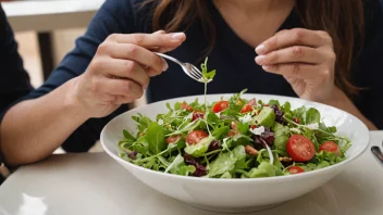 En person som sitter ved et bord på en restaurant og spiser en liten salat fra en tallerken, med et glass vann ved siden av.